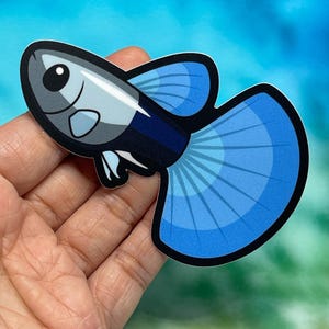 May include: A sticker of a cartoon guppy fish with a blue tail and fins. The fish has a gray body with a black outline. The sticker is held in a person's hand.