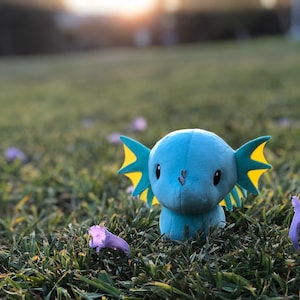 May include: A blue and yellow plush toy with large ears and big eyes sits in a field of green grass with purple flowers.