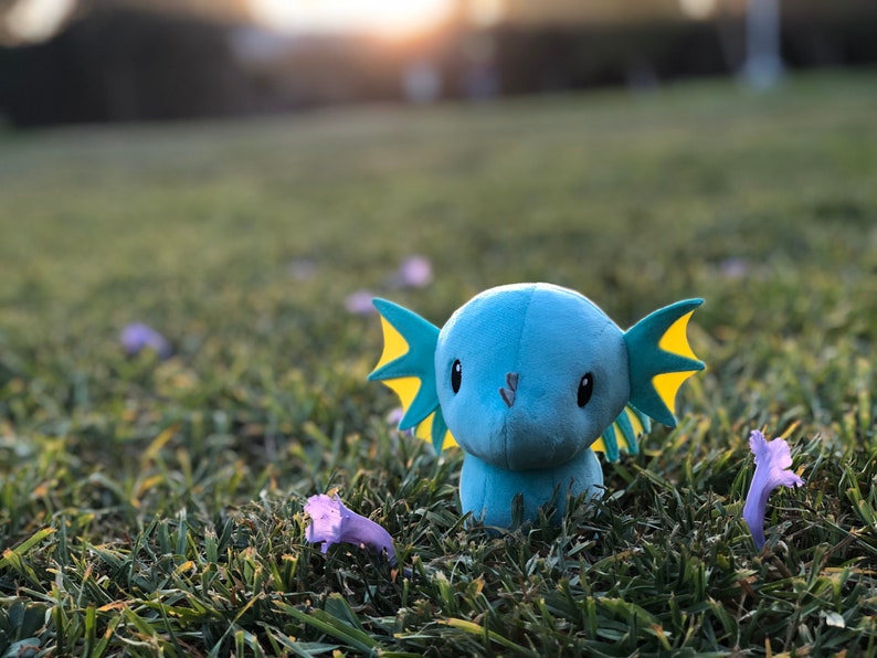May include: A blue and yellow plush toy with large ears and big eyes sits in a field of green grass with purple flowers.