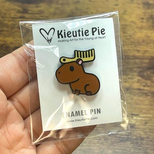 May include: A brown enamel pin featuring a cartoon moose with yellow antlers and a comb. The pin is on a white card with the text "Kieutie Pie" and "Making Art for the Young at Heart". The pin is in a clear plastic bag.