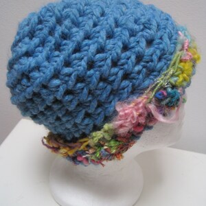 May include: A handmade blue crochet hat with a textured stitch pattern. The hat features a colorful band of yarn in shades of pink, yellow, green, and other hues. The hat is displayed on a white mannequin head.