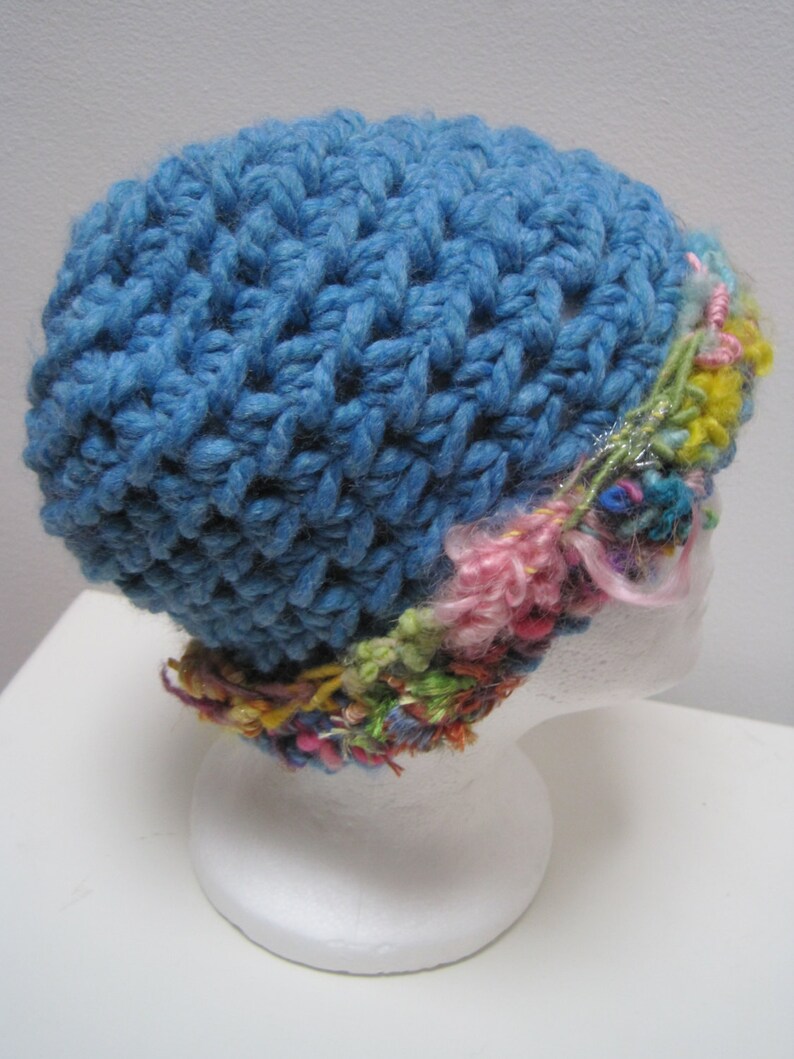 May include: A handmade blue crochet hat with a textured stitch pattern. The hat features a colorful band of yarn in shades of pink, yellow, green, and other hues. The hat is displayed on a white mannequin head.