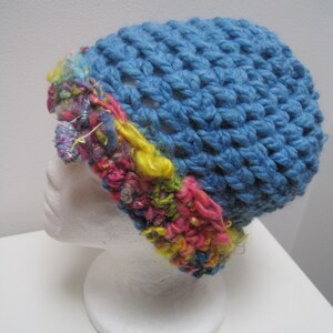 May include: A handmade blue crochet hat with a textured stitch pattern. The hat features a colorful band of mixed yarn in shades of pink, yellow, and green. The hat is displayed on a white mannequin head.