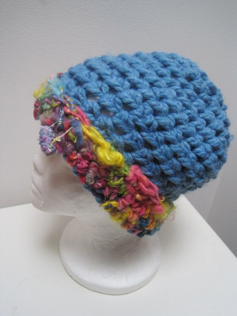 May include: A handmade blue crochet hat with a textured stitch pattern. The hat features a colorful band of mixed yarn in shades of pink, yellow, and green. The hat is displayed on a white mannequin head.