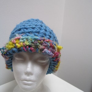May include: A blue crocheted hat with a colorful, textured brim. The brim features a mix of pink, yellow, and blue yarns, adding a vibrant contrast to the solid blue crown. The hat is displayed on a white mannequin head.