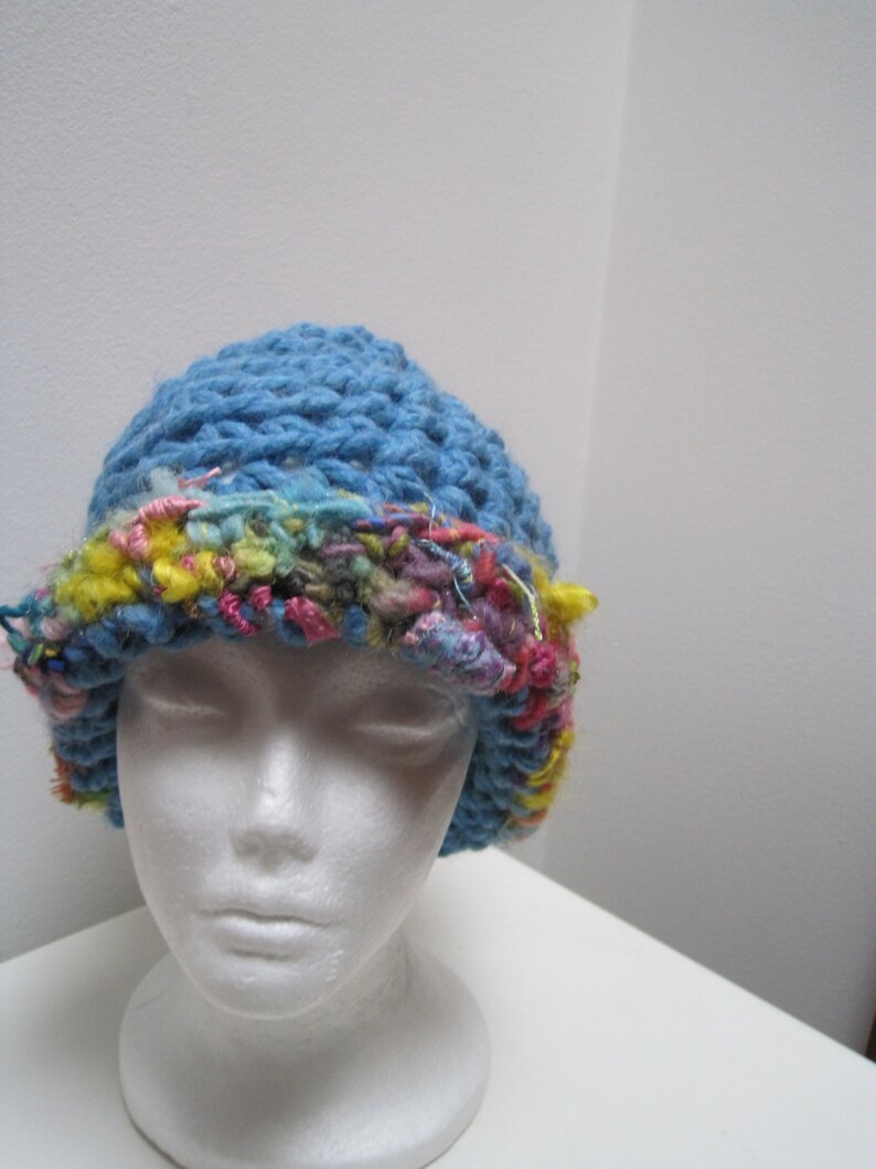 May include: A blue crocheted hat with a colorful, textured brim. The brim features a mix of pink, yellow, and blue yarns, adding a vibrant contrast to the solid blue crown. The hat is displayed on a white mannequin head.
