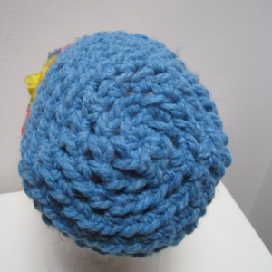 May include: A handmade blue crochet hat with a textured stitch pattern. The hat features a small, colorful floral accent on the side. The hat is displayed on a white mannequin head.