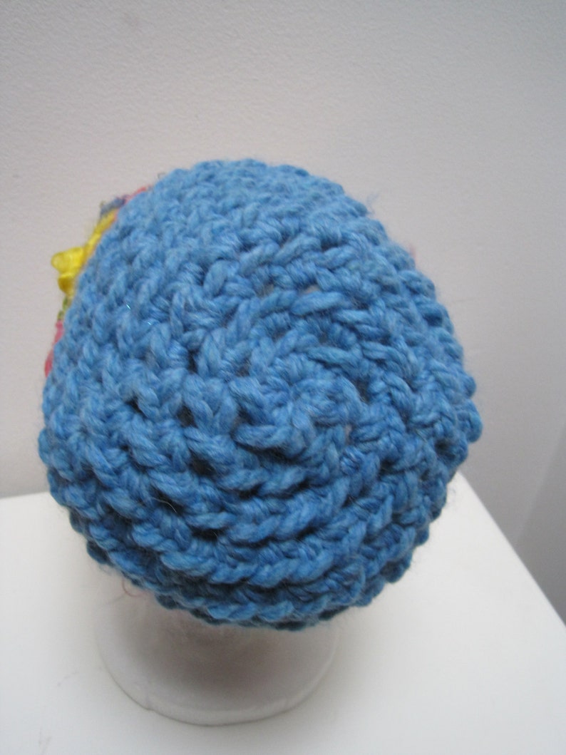 May include: A handmade blue crochet hat with a textured stitch pattern. The hat features a small, colorful floral accent on the side. The hat is displayed on a white mannequin head.