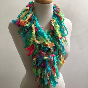 May include: A colorful, fringed scarf displayed on a mannequin. The scarf features vibrant hues of turquoise, pink, yellow, and green. The scarf's texture appears soft and flowing, with a layered design. The scarf is a fashion accessory.
