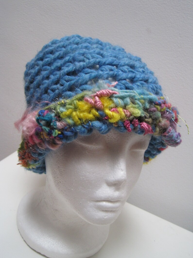 May include: A blue crochet hat with a colorful brim. The hat is made of thick yarn and features a textured stitch pattern. The brim is adorned with pink, yellow, and blue yarn, creating a vibrant contrast.