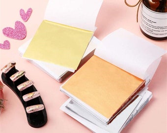 Può includere: Due pile di fogli di lamina d'oro e color rame, ciascuno con un retro di carta bianca, sono esposte. Il foglio superiore di ogni pila è sollevato, rivelando la lamina metallica. Un supporto per unghie nero con unghie dipinte è in primo piano.