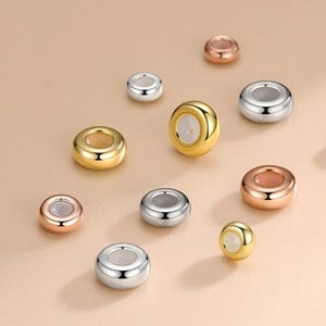 以下が含まれることがあります： 金、銀、ローズゴールドなど、さまざまな金属仕上げのジュエリービーズの詰め合わせ。ビーズは丸く、中央に穴が開いており、ネックレスやブレスレットに通すのに適しています。ビーズはニュートラルな背景に散らばっています。