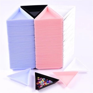 May include: A stack of 50 triangle-shaped plastic trays in white, light pink, and light blue. The trays are designed for organizing small items like beads or rhinestones. Three trays are shown in the foreground, one in each color.