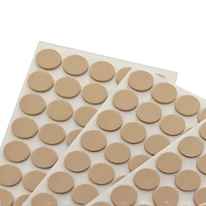Può includere: Primo piano di diversi fogli di cuscinetti adesivi circolari beige. I cuscinetti sono disposti in file ordinate su un supporto bianco, con una leggera lucentezza. I cuscinetti vengono probabilmente utilizzati per la protezione dei mobili o come paracolpi.