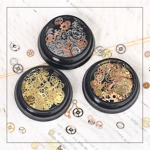 May include: Three round containers filled with assorted gold, silver, and copper gears and clock parts. The containers are black with a clear lid.