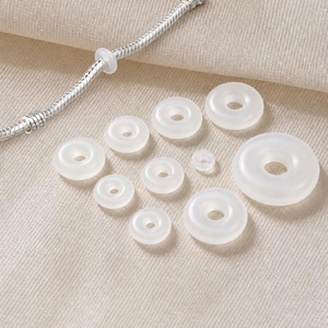 Peut inclure: Un ensemble de dix anneaux d'espacement en silicone blanc translucide de tailles variées disposés en groupe, accompagné d'un bracelet en chaîne serpent argenté comportant un anneau en silicone assorti enfilé sur la chaîne.
