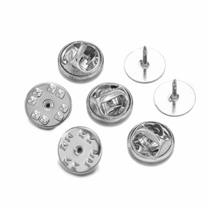 Może przedstawiać: Sześć srebrnych metalowych spinek do przypinania odznak lub innych przedmiotów do odzieży. Spinki mają okrągły kształt o średnicy około 2,5 cm.