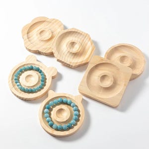 May include: A collection of wooden jewelry display trays in various shapes, including round, square, and flower-shaped. Two trays hold blue beaded bracelets. The trays are made of light-colored wood.