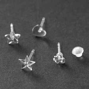 May include: A collection of clear, plastic stud earrings in various shapes, including a star, a heart, a flower, and a floral design. The earrings are displayed on a dark gray surface.