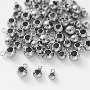 May include: A collection of shiny, silver-colored metal beads with a hollow center and a small loop for stringing. The beads vary in size, with a few larger ones and many smaller ones scattered throughout.