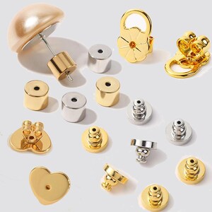 Puede incluir: Una variedad de componentes de pendientes en tonos dorados y plateados. Incluye varios cierres, postes y elementos decorativos como una flor y un respaldo en forma de corazón. Los componentes se muestran sobre un fondo claro.