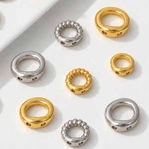 May include: A collection of circular metal jewelry components in gold and silver tones. The set includes smooth rings and textured rings with beaded edges, each featuring two small holes on the side for attachment, arranged on a white surface.