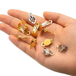 May include: A collection of gold and silver-colored clip-on earring findings. The findings are in various shapes and sizes, with a hinged clip mechanism for attaching to the ear.