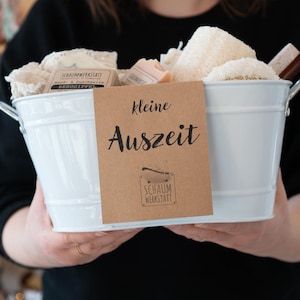 Könnte beinhalten: Ein weißer Metalleimer gefüllt mit Badeprodukten, darunter Seife, Waschlappen und ein Luffa. Ein braunes Papieretikett mit dem Text "kleine Auszeit" und "Schaum Werkstatt" ist am Eimer befestigt.