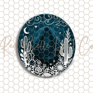 May include: A round, dark teal design featuring a cowboy hat silhouette. White floral and cactus illustrations frame the hat, with a crescent moon and stars at the top. The words "Belle & Co" are written across the hat.