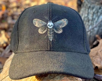 May include: Black baseball cap with a white and gray moth with a skull design on the front.