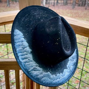 May include: A black fedora hat with a white cloud design and gold glitter accents. The hat is decorated with a white cloud pattern that resembles a starry night sky.