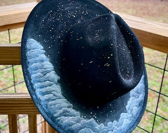 May include: A black fedora hat with a white cloud design and gold glitter accents. The hat is decorated with a white cloud pattern that resembles a starry night sky.