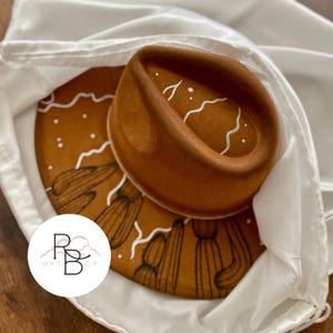 May include: A brown fedora hat with white painted accents and a matching brim featuring a cactus design. The hat is nestled in a white cloth. The logo "RB HAT CO." is visible in the lower left corner.