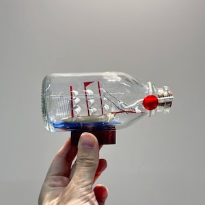 Può includere: Una bottiglia di vetro trasparente con una nave a vela in miniatura all'interno. La nave ha vele rosse ed è posta su una base blu. Il tappo è adornato da una perla rossa. La bottiglia misura circa 15 cm di lunghezza.