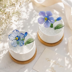 Può includere: Due sculture di fiori in vetro in vasi di ceramica bianca, ciascuna su un sottobicchiere di legno. I fiori sono azzurri con centri gialli e foglie verdi. I vasi sono riempiti di ciottoli bianchi. È visibile un biglietto di auguri con la scritta "Especially for you".