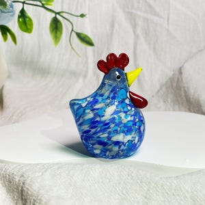 May include: A blue and white glass chicken figurine with a red comb and yellow beak. The figurine is sitting on a white surface.