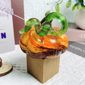 Puede incluir: Una calabaza de vidrio con un tallo de espiral verde, sentada sobre una base de madera. La calabaza es naranja y marr&oacute;n con un efecto marmoleado.
