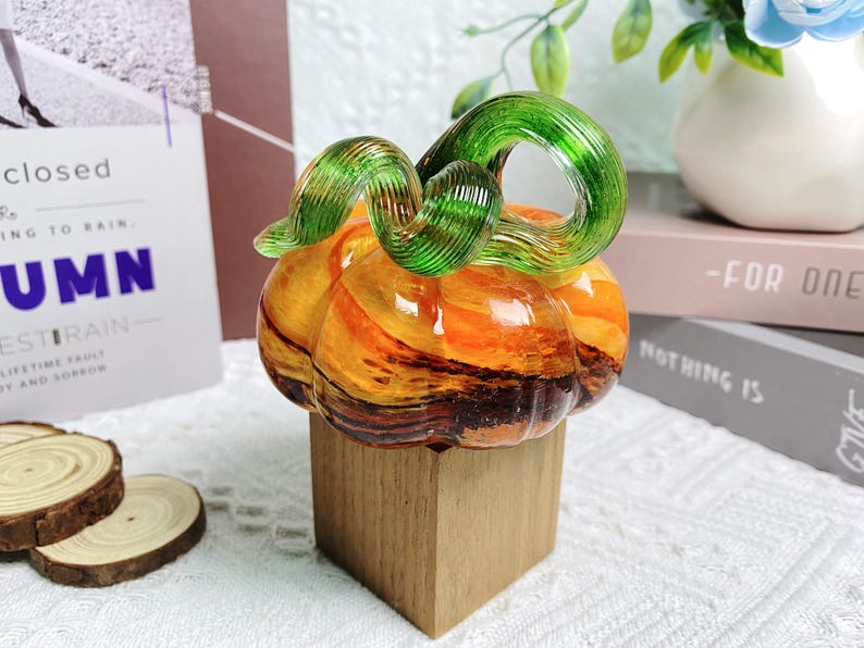 Puede incluir: Una calabaza de vidrio con un tallo de espiral verde, sentada sobre una base de madera. La calabaza es naranja y marr&oacute;n con un efecto marmoleado.