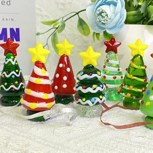 May include: Seven glass Christmas tree ornaments in various colors and designs. The ornaments are decorated with white, red, green, and yellow paint. The ornaments are arranged on a white surface.