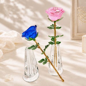 Può includere: Due rose di vetro, una rosa e una blu, ciascuna con uno stelo dorato e foglie verdi. Le rose sono esposte in due vasi di vetro trasparente sfaccettato. I petali delle rose hanno un aspetto traslucido e cristallino.
