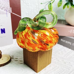 Puede incluir: Una figurita de calabaza de vidrio con un tallo verde y remolinos naranja y amarillo. La calabaza est&aacute; sentada sobre una base de madera.