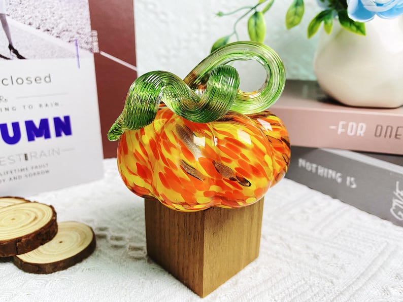 Puede incluir: Una figurita de calabaza de vidrio con un tallo verde y remolinos naranja y amarillo. La calabaza est&aacute; sentada sobre una base de madera.