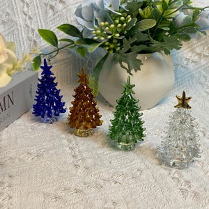 Puede incluir: Cuatro adornos de árbol de Navidad de vidrio en diferentes colores. Los adornos son azules, dorados, verdes y transparentes con una estrella dorada en la parte superior. Los adornos están sobre una superficie blanca.