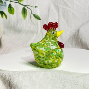 May include: A green glass chicken figurine with white and yellow speckles. The chicken has a red comb and a yellow beak.