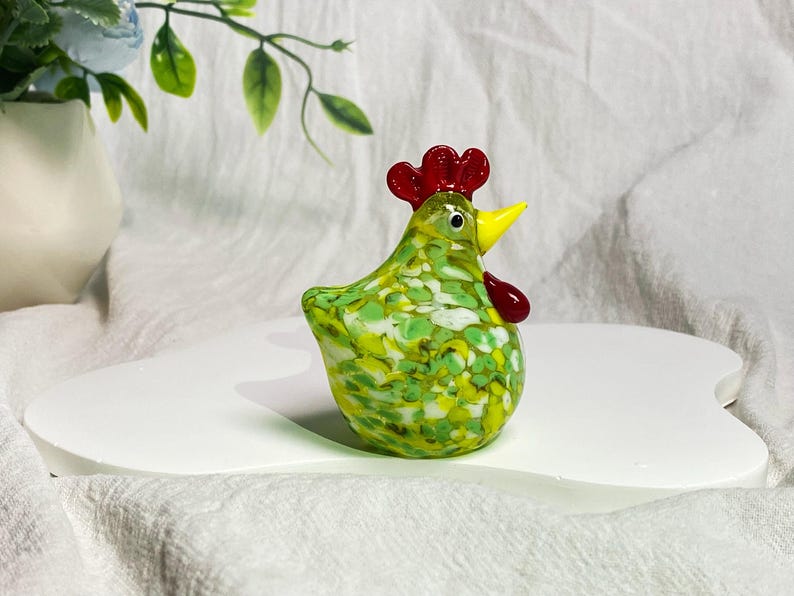May include: A green glass chicken figurine with white and yellow speckles. The chicken has a red comb and a yellow beak.