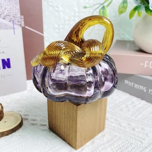 Puede incluir: Una calabaza de vidrio con un dise&ntilde;o de espiral en morado y dorado, sentada sobre una base de madera.