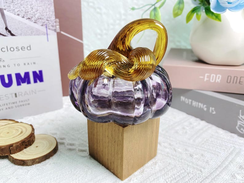 Puede incluir: Una calabaza de vidrio con un dise&ntilde;o de espiral en morado y dorado, sentada sobre una base de madera.