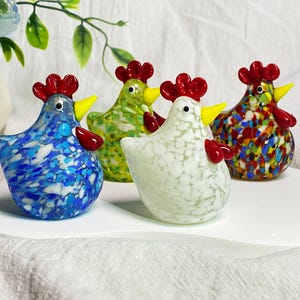 May include: Four glass chickens in different colors and patterns. The chickens are standing on a white surface. The chickens are all facing forward and have red combs and wattles.