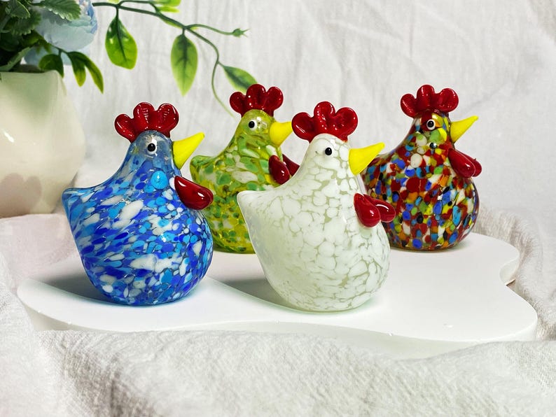 May include: Four glass chickens in different colors and patterns. The chickens are standing on a white surface. The chickens are all facing forward and have red combs and wattles.