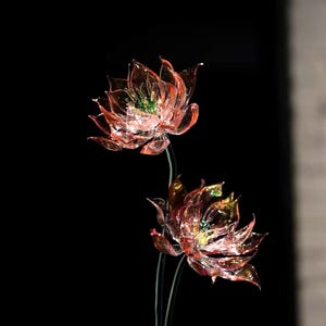 Puede incluir: Dos flores de loto artificiales con pétalos rojos y dorados translúcidos y centros verdes. Las flores están sobre tallos finos y oscuros, sobre un fondo negro. Los pétalos tienen una apariencia delicada, como de cristal.